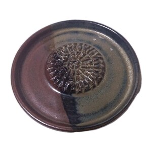 May include: A handmade ceramic garlic grater with a textured center for grating. The plate is round with a dark brown and blue-green glaze. The grater has a small handle on the side.