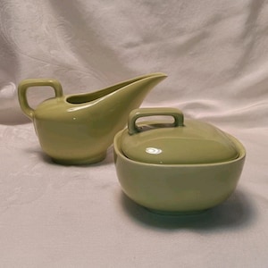 May include: A light green ceramic creamer pitcher with a curved handle and a pointed spout, paired with a matching lidded sugar bowl featuring a square handle on the lid. Both pieces have a smooth, solid-colored finish and a rounded base design.