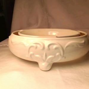 May include: A cream-colored ceramic serving bowl with a low, wide profile and a smooth, glossy finish. The exterior features raised, symmetrical scrollwork patterns and the bowl is supported by three short, integrated feet at the base.