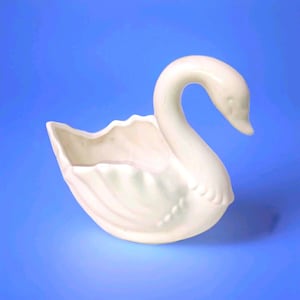May include: A white ceramic swan planter with a curved neck and open basin. The swan has detailed feather and wing designs. The planter is set against a blue background.