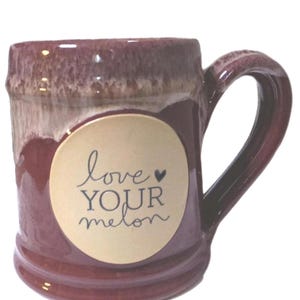 Puede incluir: Una taza de cerámica granate con un asa redondeada. La taza presenta una etiqueta circular de color crema con las palabras "love YOUR melon" en un estilo manuscrito. La taza tiene un acabado brillante y un borde texturizado.