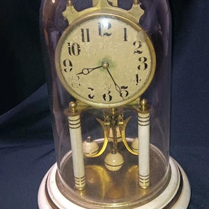May include: An antique clock with a cream-colored face, black numerals, and ornate gold-colored hands. The clock is encased in a clear glass dome, resting on a white and gold base. The clock's pendulum and decorative elements are visible.