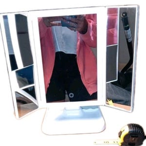 Puede incluir: Un espejo de tocador blanco de tres pliegues con una base rectangular. El espejo tiene una luz incorporada y está abierto, reflejando a una persona que lleva una chaqueta rosa y pantalones cortos negros. Una cinta métrica amarilla y negra está en primer plano.