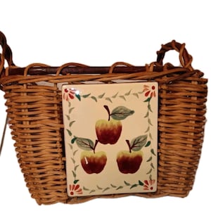 Op de afbeelding: Wicker mand met een rechthoekige keramische tegel met drie rode en gele appels met groene bladeren. De mand heeft twee handvatten en een donkerbruine houten balk.