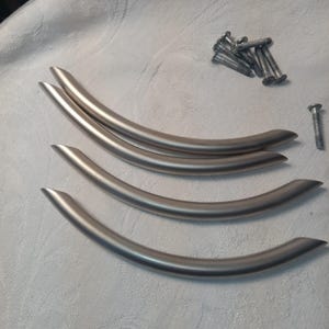 Op de afbeelding: Vier kastgrepen van geborsteld nikkel met een gebogen, boogvormig ontwerp. Elke handgreep heeft een gladde, metalen afwerking en taps toelopende uiteinden. Een kleine set bijpassende zilverkleurige metalen montageschroeven is inbegrepen.