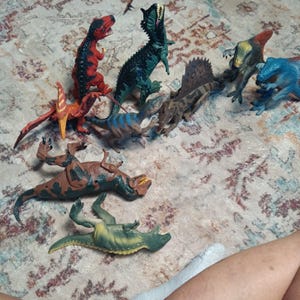May include: A collection of plastic dinosaur figurines in various poses and colors, including a red and black bipedal dinosaur, a green spiky dinosaur, a brown and blue striped dinosaur, a brown spined dinosaur, and a green and yellow dinosaur.