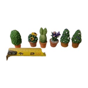 Puede incluir: Pequeñas plantas en macetas, incluyendo girasoles, una planta con forma de conejo y plantas con flores, dispuestas en fila. Las plantas están en pequeñas macetas de terracota. La altura es de unos 5 cm.