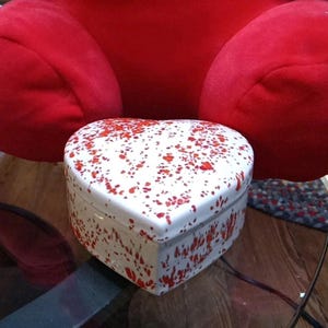 May include: A heart-shaped trinket box with a white base and red splatter design. The box is ceramic and has a lid. The background features a large red plush object.