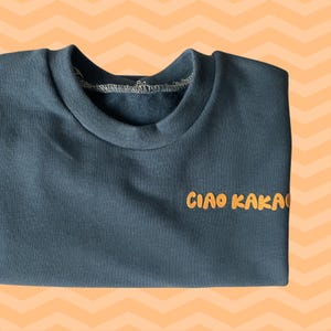 Op de afbeelding: Een donkerblauwgroen sweatshirt met de tekst "CIAO KAKA" in oranje letters. De crew neck sweatshirt is gemaakt van een zacht materiaal en wordt gepresenteerd op een oranje en witte chevron achtergrond.