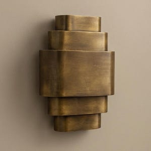 Könnte beinhalten: Eine messingfarbene Wandleuchte mit einem geschichteten, geometrischen Design. Die Leuchte hat eine Reihe von rechteckigen Formen, die übereinander gestapelt sind und ein gestuftes Aussehen erzeugen. Die Leuchte ist an einer neutral getönten Wand befestigt.