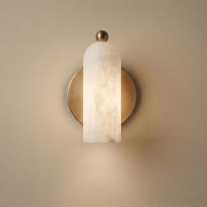 Puede incluir: Aplique de pared moderno con una pantalla rectangular de alabastro blanco y una placa trasera circular de color latón. La lámpara está rematada con una pequeña esfera de color latón. Montado en una pared beige.