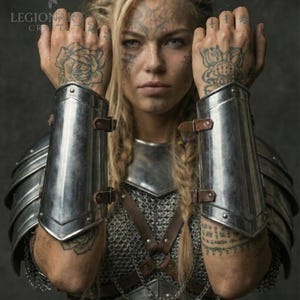 May include: A person wearing silver metal arm guards and chest armour. The person has blonde hair in braids and tattoos on their face and hands. The armour is detailed with leather straps and chainmail.