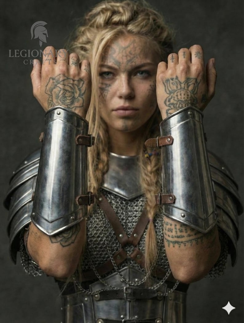 May include: A person wearing silver metal arm guards and chest armour. The person has blonde hair in braids and tattoos on their face and hands. The armour is detailed with leather straps and chainmail.
