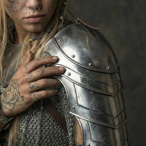 May include: A silver shoulder pauldron with riveted plates, part of a suit of armour. The pauldron is worn over chainmail. A leather strap is visible. The image shows a close-up of the shoulder and upper chest.