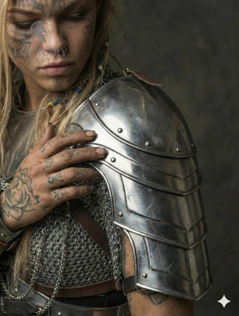 May include: A silver shoulder pauldron with riveted plates, part of a suit of armour. The pauldron is worn over chainmail. A leather strap is visible. The image shows a close-up of the shoulder and upper chest.