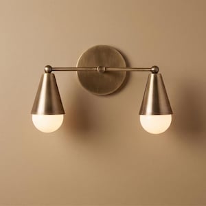 Pode incluir: Arandela de parede em latão com duas cúpulas cônicas, cada uma com uma lâmpada redonda. A luminária é montada em uma placa traseira circular e uma barra horizontal. Ideal para iluminação interior.