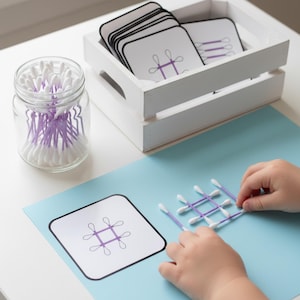 May include: A clear glass jar filled with white cotton swabs with purple stems. A white wooden crate holds cards with purple line drawings. A child is arranging cotton swabs on a blue surface, matching the card designs.