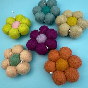 May include: Six felt flower-shaped decorations in various colours, including green, pink, purple, and orange. Each flower is made of multiple felt balls and has a small white loop for hanging. The flowers are arranged on a light blue background.