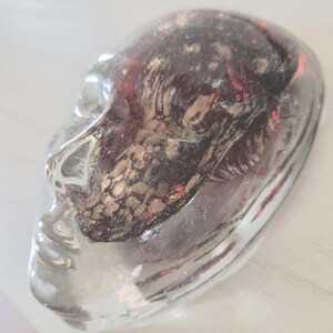May include: A clear, molded resin sculpture shaped like a human face in profile. The interior contains a dark, textured layer with metallic gold and bronze flecks, along with small red accents embedded within the translucent, glossy material.
