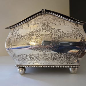 Könnte beinhalten: Eine silberfarbene Metallbox mit Klappdeckel, die auf der Vorderseite mit detaillierten Blumen- und Schleifengravuren verziert ist. Der Behälter hat eine geschwungene Form, einen Perlenrand an der Oberkante und steht auf vier kleinen, kugelförmigen Füßen.