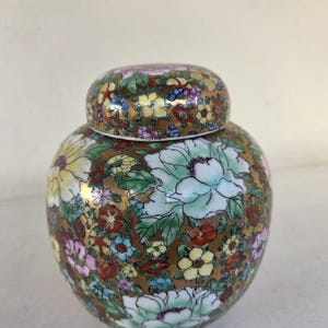 Pode incluir: Um pote de gengibre de cerâmica redonda com tampa correspondente, apresentando um padrão floral denso e colorido de flores brancas, amarelas, rosas e vermelhas contra um fundo dourado metálico. O vaso tem um corpo bulboso e acabamento liso.