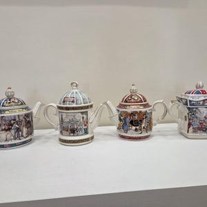 Op de afbeelding: Vier keramische theepotten met gedetailleerde illustraties van stadsgezichten en bezienswaardigheden. Elke theepot heeft een uniek ontwerp voor het deksel, het handvat en de tuit, met kleurrijke beschilderde motieven, gouden accenten en patronen.