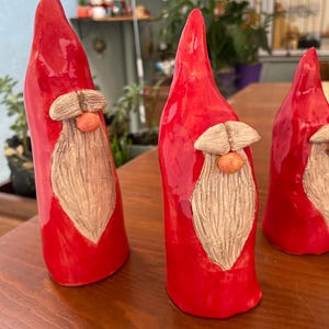 May include: Three ceramic gnome figurines with bright red conical hats and bodies. Each gnome has a tan beard, a small orange nose, and a tan mustache. The figurines are displayed on a wooden surface.