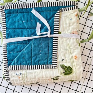May include: A square, quilted baby blanket with a teal center and a frog and lily pad pattern on the exterior. The blanket is bound with black and white striped fabric and tied with a white ribbon.