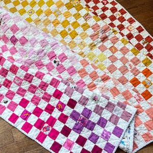 May include: A checkered quilt with a rainbow gradient of colors, starting with yellow and ending with pink. The quilt is made of squares of different fabrics, each with a unique pattern. The quilt is folded in half and lying on a wooden floor.
