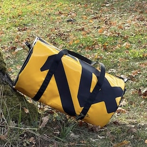 Può includere: Una borsa da viaggio gialla e nera è appoggiata a terra vicino a un albero. La borsa ha manici neri e un grande disegno grafico nero sul lato. Lo sfondo presenta erba, foglie cadute e tronchi d'albero.