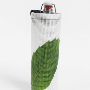 Peut inclure: Porte-bouteille blanc avec un grand motif de feuille verte. Le support a une texture perforée et contient une bouteille en verre avec un bouchon rouge et blanc. Une sangle noire est attachée en haut.