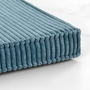 A close-up view of a textured, blue-green fabric with a ribbed or striped pattern, resting on a white surface.