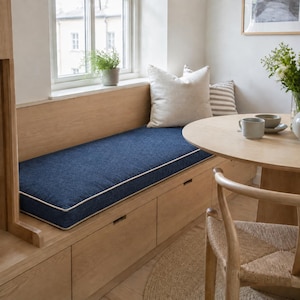 May include: A built-in wooden bench featuring a long, rectangular navy blue cushion with white piping. The bench includes integrated storage drawers underneath and is topped with a large, square, off-white throw pillow and a smaller striped pillow.