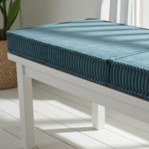 A white bench with a blue cushion on top, situated in a room with a window and a plant.