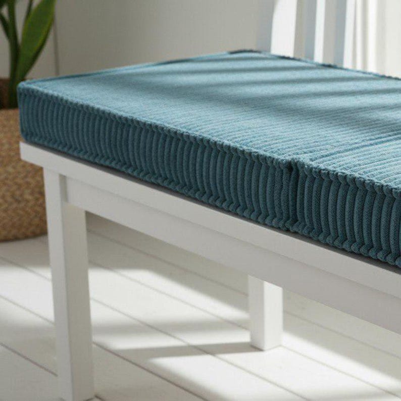 A white bench with a blue cushion on top, situated in a room with a window and a plant.