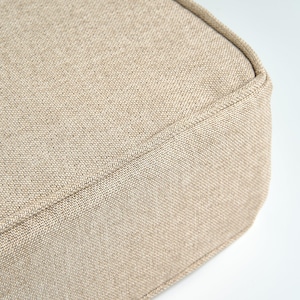 May include: Close-up of a beige fabric cushion corner. The fabric has a textured appearance with visible woven threads. The edges are neatly sewn with a matching thread, creating a clean, finished look. The neutral colour suggests versatility for various home decor styles.