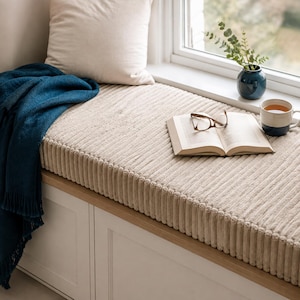 May include: A beige corduroy window seat cushion with a textured ribbed surface. The cushion is topped with an open book, brown-rimmed reading glasses, a dark blue ceramic mug with a light-colored rim, and a dark blue throw blanket.