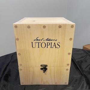 May include: A light-colored wooden box with the words "Sam Adams Utopias" in black script. The box has a metal clasp and is constructed with visible wood joints and small, round fasteners.