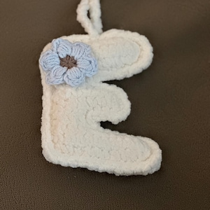 May include: A white, fluffy, crocheted letter "E" keychain with a light blue and brown flower accent. The keychain has a gold-colored clasp for easy attachment. The soft texture and floral detail make it a charming accessory.