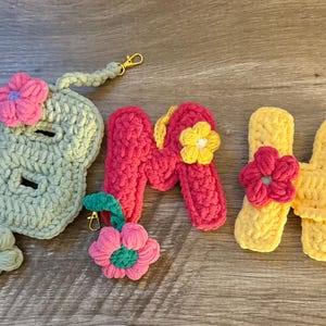 May include: Crocheted accessories in the image include a light green case with a pink flower, the letters "M" and "H", and a flower-shaped keychain. The letters are pink and yellow, each adorned with a flower. All items have gold-colored clasps.