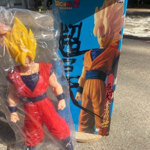 May include: A Dragon Ball Z Son Goku action figure in a plastic bag, next to its box. The figure is red and blue, with yellow spiky hair. The box is blue with Japanese text and an image of Goku. The box states the figure is 36 cm tall.