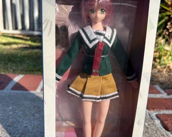 May include: A doll in a box with pink hair, green eyes, and a school uniform. The uniform is green with a white collar and a red tie. The box reads "Natural Body is used" and "Doll Collection".