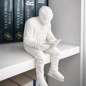 Könnte beinhalten: Eine weiße Skulptur eines Eishockeyspielers, der ein Buch liest und auf einem Regal sitzt. Die Figur trägt Helm, Trikot, Hose, Handschuhe und Schlittschuhe. Dunkelblaue Bücher mit silbernen Buchstaben befinden sich dahinter.