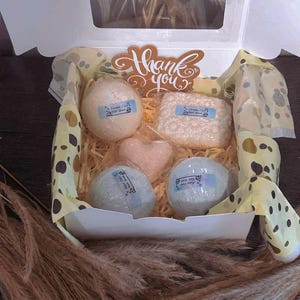Puede incluir: Una caja de regalo abierta con bombas de baño y un objeto en forma de corazón, sobre papel decorativo. La caja incluye un mensaje de "Gracias". Las bombas de baño son de varios colores y formas, con etiquetas.