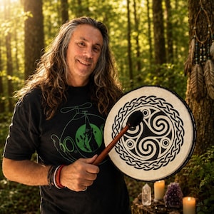 May include: A round frame drum featuring a black triskelion symbol and an intricate Celtic knot border on the drumhead. The drum is held with a wooden mallet that has a black padded head. The drumhead is light-colored with a dark rim.
