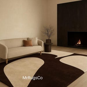 May include: A modern living room featuring a beige sofa with brown accent pillows, a dark brown rug with a beige abstract design, and a fireplace. A small side table holds a decorative plant. The room has a neutral color palette.