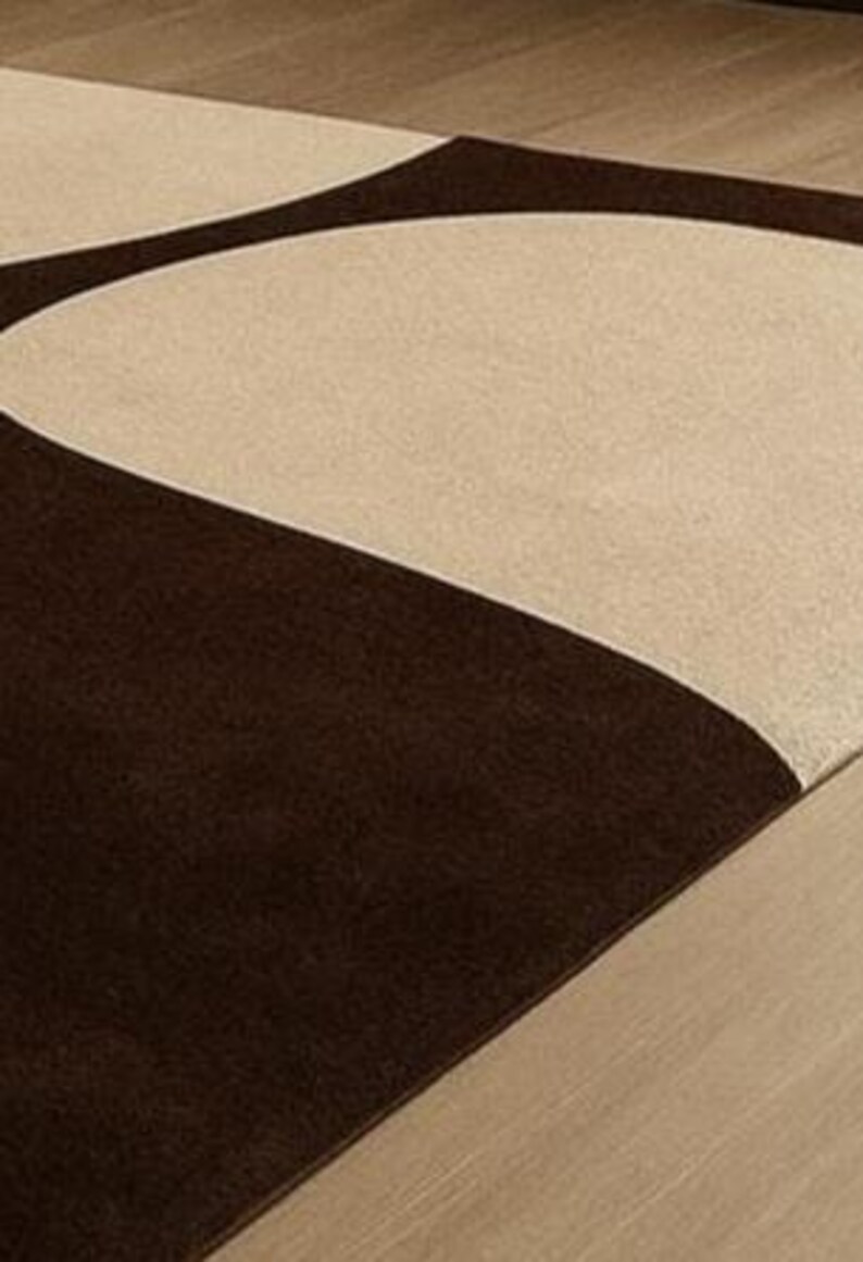 May include: A modern area rug featuring a geometric design with a combination of brown and beige colors. The rug's pattern includes curved shapes, creating a contemporary aesthetic. The rug is placed on a wooden floor.