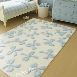 Può includere: Un tappeto color crema con un motivo a fiocchi azzurri. Il tappeto si trova nella cameretta di un bambino, con un letto, un comò e un camion giocattolo visibili. Il tappeto misura circa 152 cm per 229 cm.