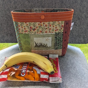 May include: A patchwork fabric tote bag with an orange trim and button closure. The bag contains a banana, a bag of Cheez-Its, and a caramel candy bar. The bag features a cat design.