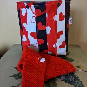 May include: A red and black striped fabric bag with white hearts printed on it. The bag has a drawstring closure and is sitting on a table with a red knitted scarf.
